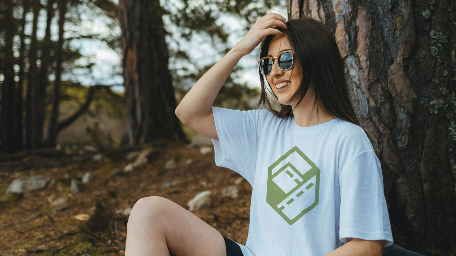 A woman in sunglasses sitting against a tree in a forest, wearing a white t-shirt with the green ExploreQuest logo
