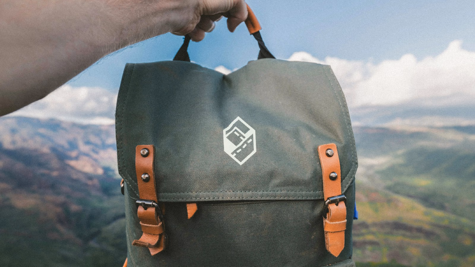 A hand holding up a dark green canvas backpack printed with the white ExploreQuest logo, mountains blurred behind