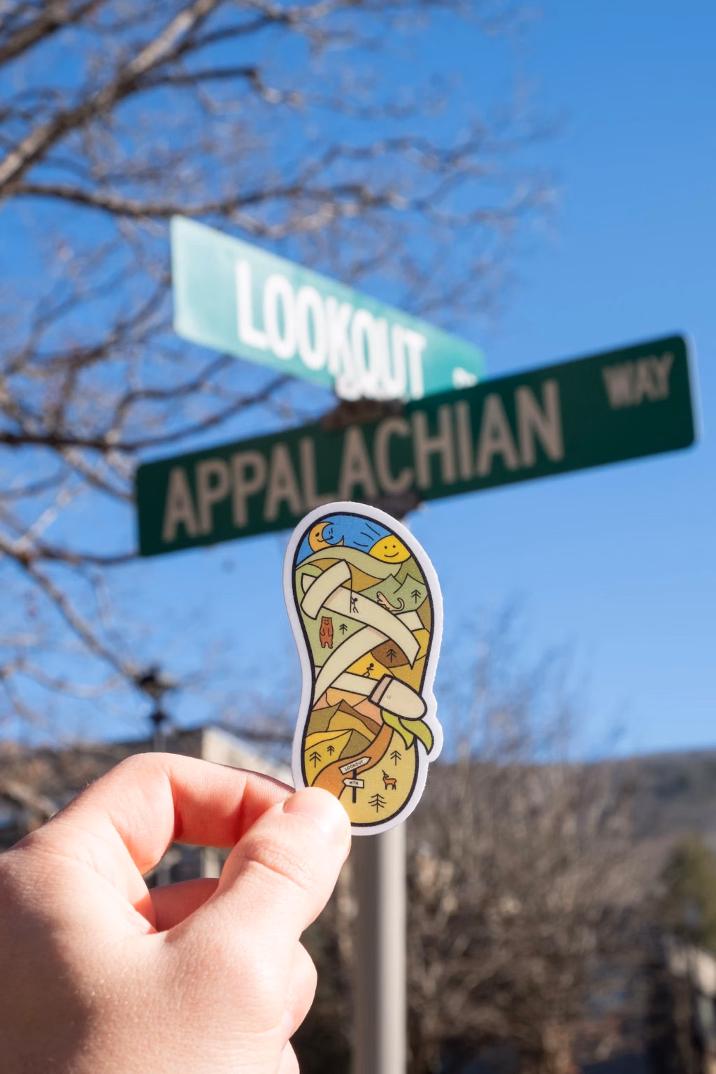 Photo of a hand holding the Chaco sandal sticker below "Lookout Dr" and "Appalachian Way" street signs against a blue sky