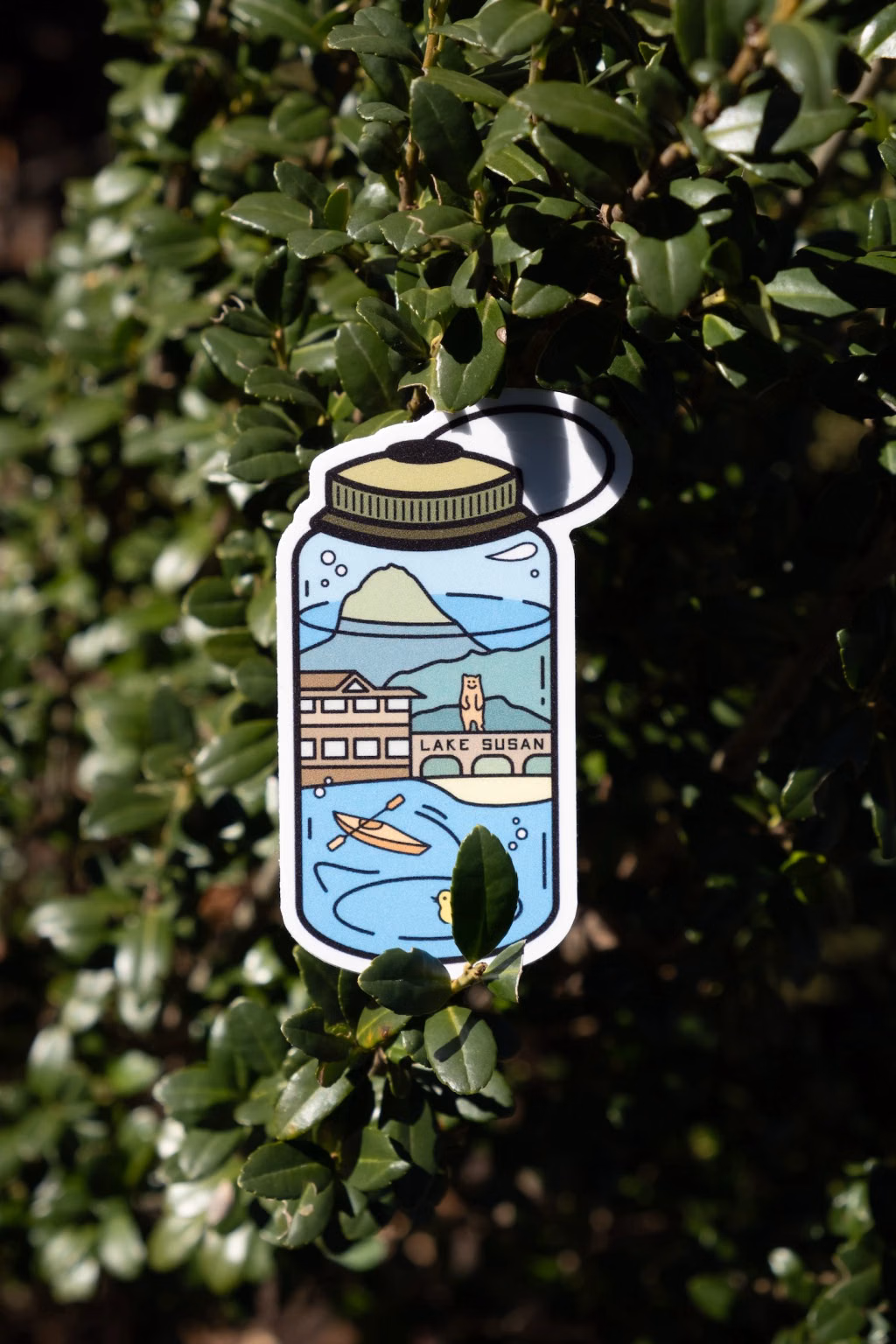 Photo of the die-cut Lake Susan Nalgene bottle sticker resting on sunlit green shrub leaves