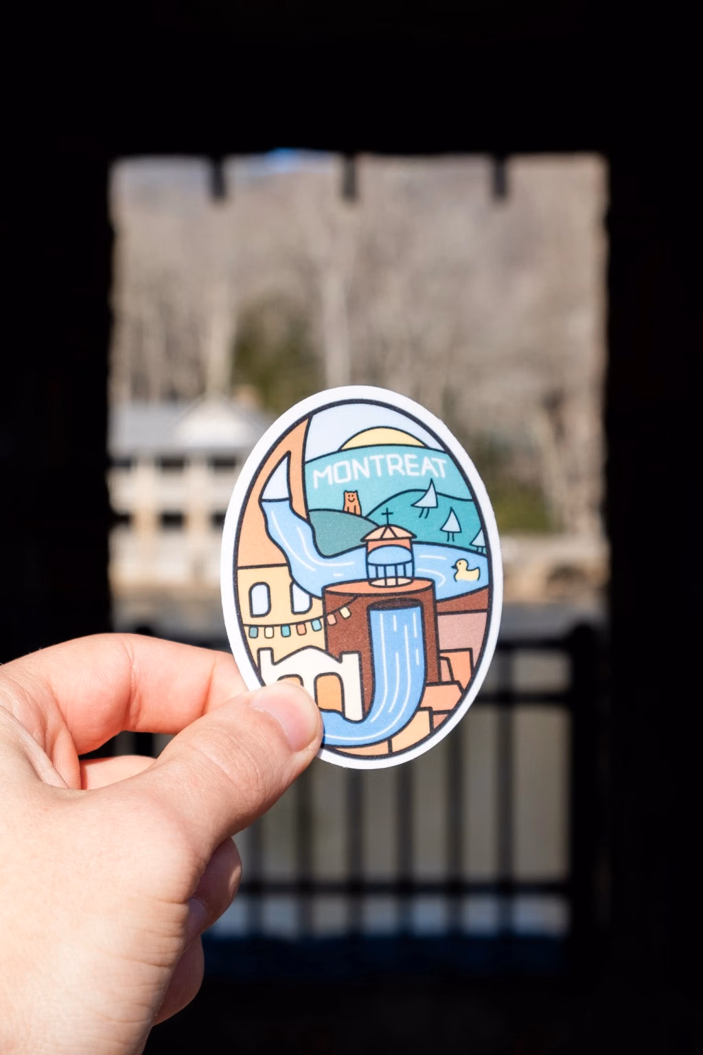 Photo of a hand holding the Montreat oval sticker in front of a stone-arch window overlooking campus