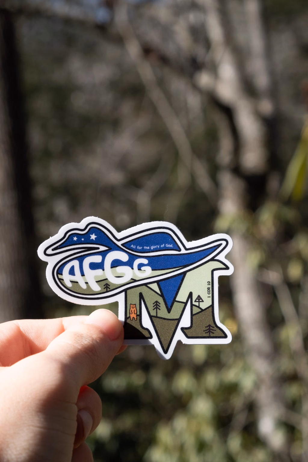 Photo of a hand holding the AFGG mountain sticker in front of a blurred winter forest