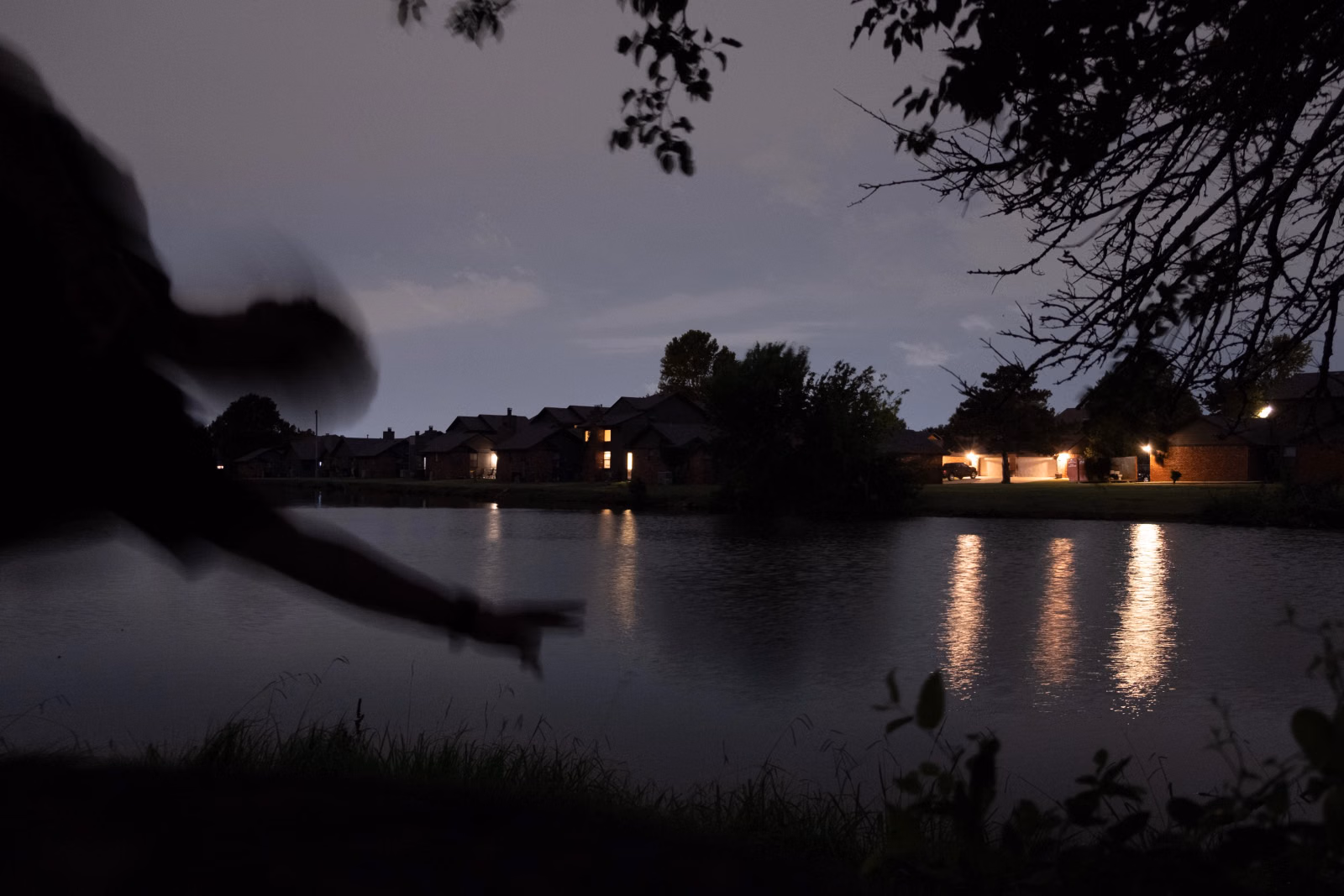 A dark silhouette of a person leaning out over a pond at twilight, one arm reaching toward the water, as warm lights from houses across the lake streak in reflections on the surface